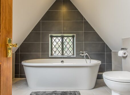 Bathroom door open showing a free standing bath against an apex wall with a window in it and wall tiles that are grey. There is a grey rug on white tiles and the celing and walls are white.