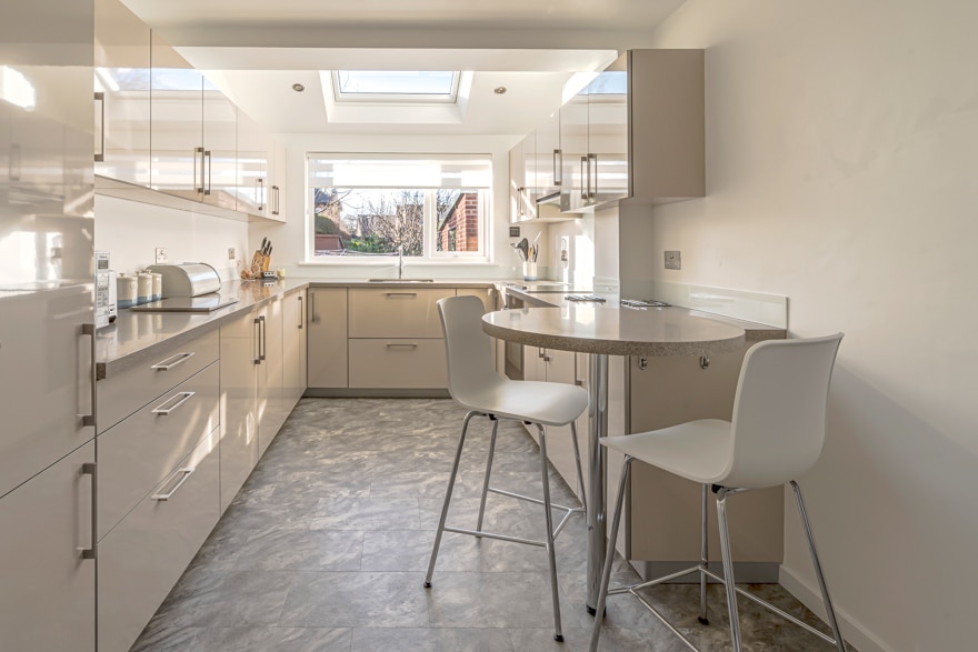 A cream kitchen in a garage conversion with units either side and a small round table with two chairs at the end. There is a roof light ans a winodw loooking out onto the road.