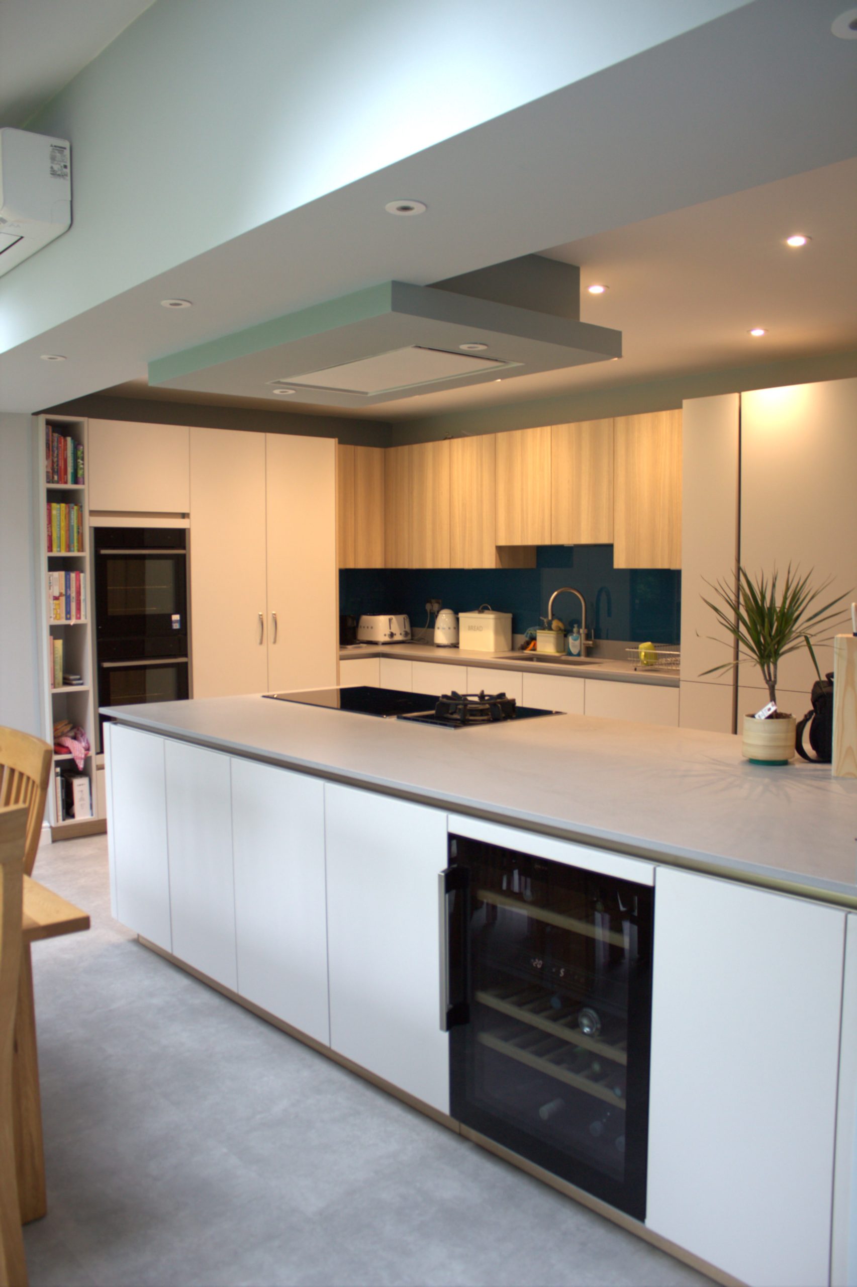 A modern kitchen with dark blue glass splashback, light oak cabinetry and modern, black appliances. A white island is in the middle with light grey countertops and a wine fridge.