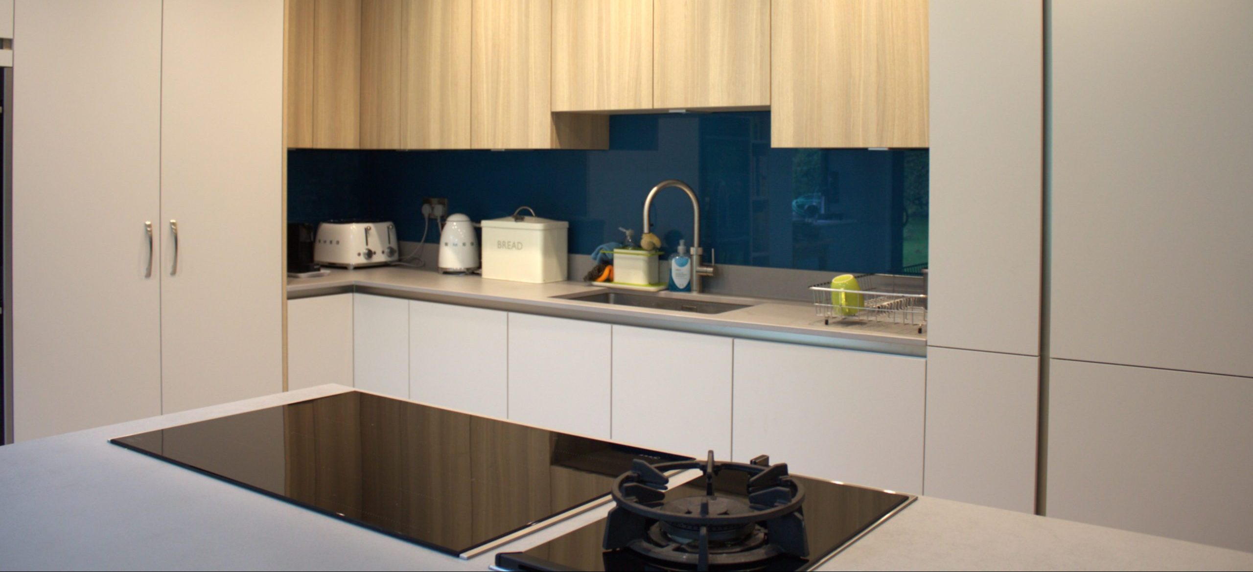 A modern kitchen with dark blue glass splashback, light oak cabinetry and modern, black appliances